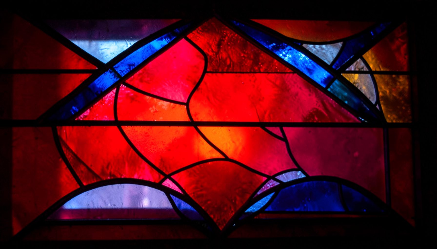 A stained-glass composition with bold leaded segments, rich jewel tones of ruby, sapphire, and amber, illuminated by soft light.