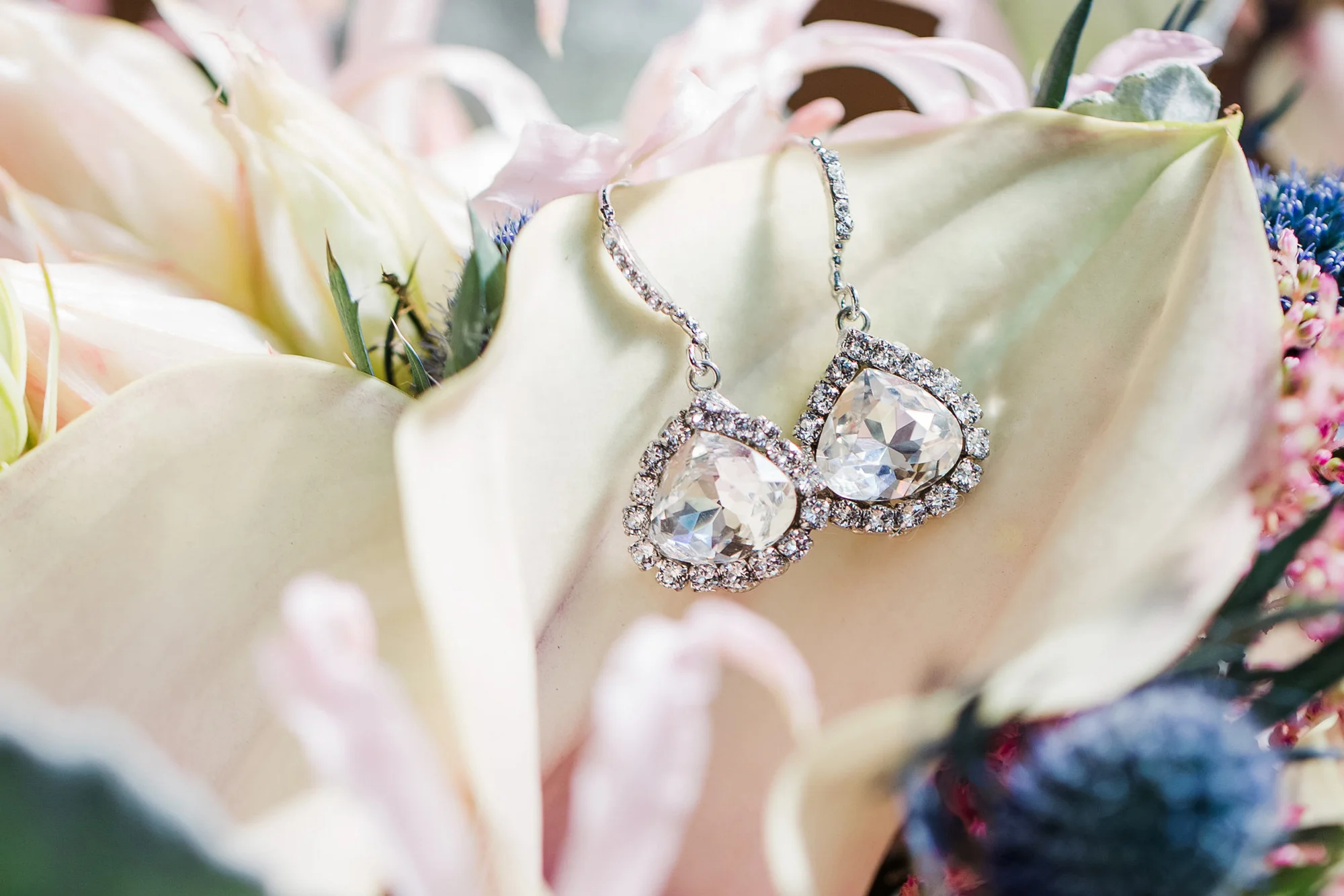 Close-up of teardrop crystal earrings resting on pale flower petals, white and pink tones.