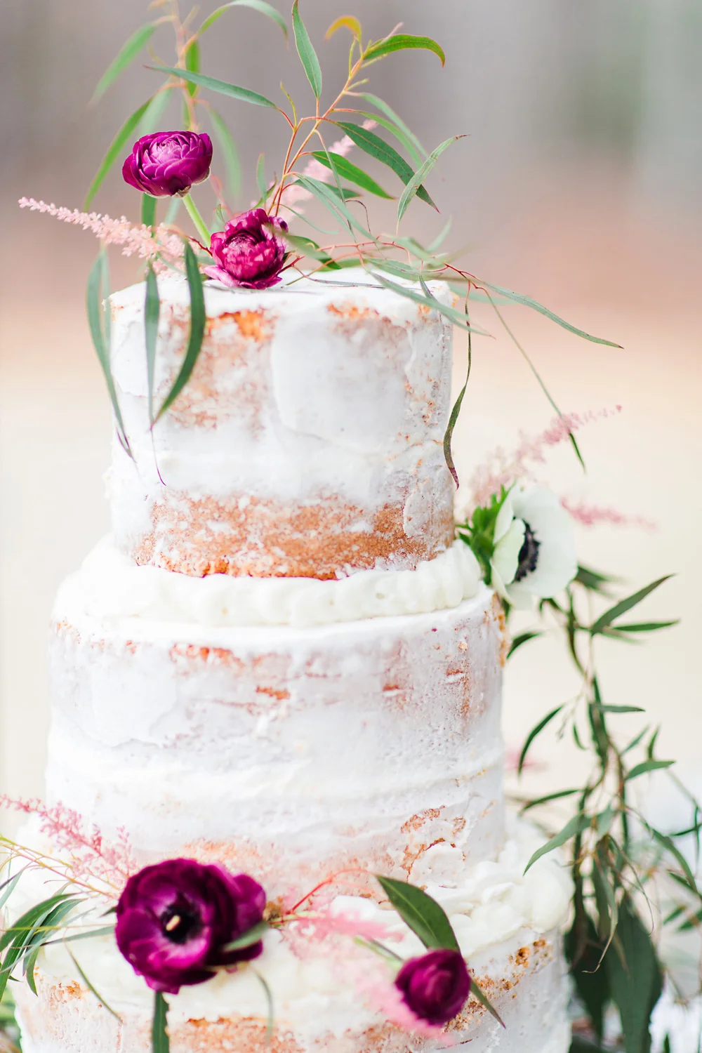 Close-up of a white textured tiered cake adorned with dark pink flowers, small white blooms, and trailing greenery.